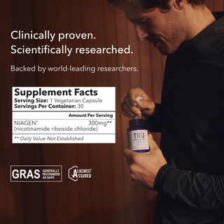 A person holding a Tru Niagen bottle, showing the Supplement Facts panel and certification logos for “Generally Recognized As Safe” and “Alkemist Assured.” The label indicates a serving size of one vegetarian capsule, with 30 servings per container. Each serving provides 300 mg of Niagen.