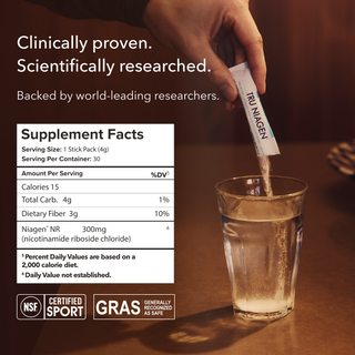 Hand holding a supplement stick pack labeled Tru Niagen over a glass of water with showing the Supplement Facts panel and certification logos for “Generally Recognized As Safe” and “Alkemist Assured.”