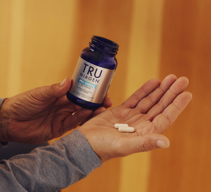 Person holding a bottle of Tru Niagen and two capsules in their hand against a blurred background.